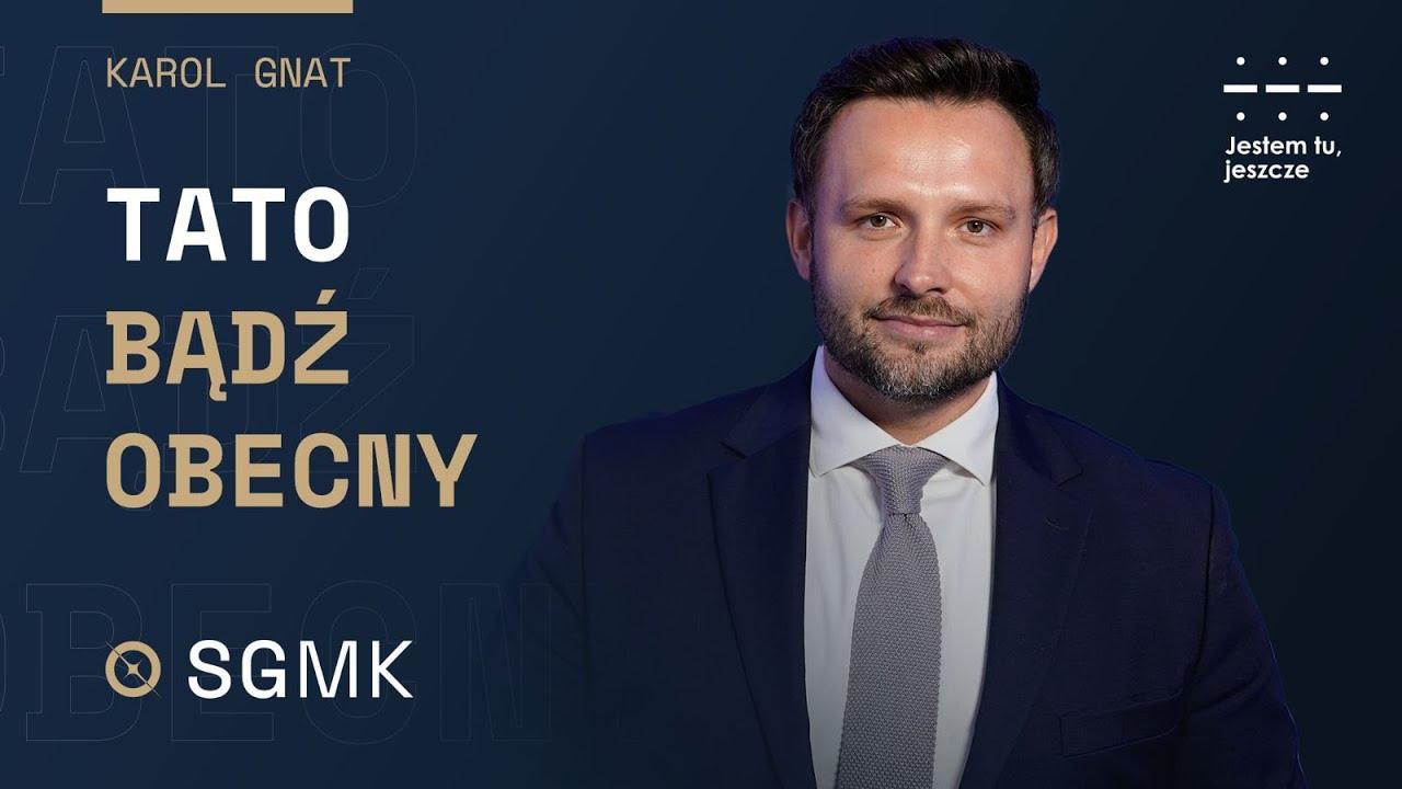 Miniatura filmu. Karol Gnat, uśmiechnięty mężczyzna w garniturze, na ciemnym tle. Po lewej stronie widnieje tytuł "Tato, bądź obecny" oraz logo SGMK. W prawym górnym rogu hasło "Jestem tu, jeszcze".